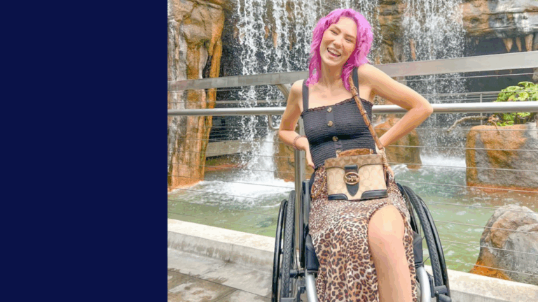 A young woman with bright pink hair smiles while sitting in her wheelchair in front of a waterfall. She wears a black top, leopard print skirt, and carries a crossbody purse.