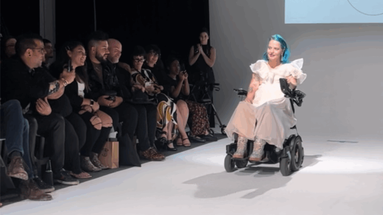 A woman with blue hair, wearing a white dress and boots, rolls down the runway in a motorized wheelchair as part of Chelsies Story, while an audience seated on both sides watches and takes photos.