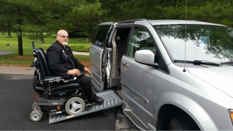 Johns Story: A man in a wheelchair uses a metal ramp to board a silver accessible van parked on a paved road, surrounded by green trees and grass.