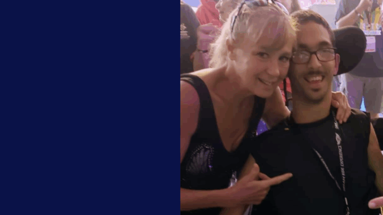 A woman smiles and leans in towards a man using a wheelchair, both posing happily at an event with people and colorful lights in the background. The left side of the image features a solid dark blue block, capturing a moment from Darlenes Story.