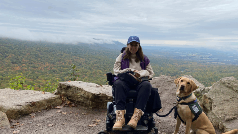 Rachels Story: A woman in a wheelchair sits on a mountain overlook with her service dog beside her. She wears a blue cap and boots. The background reveals a misty, tree-covered valley under drifting clouds.