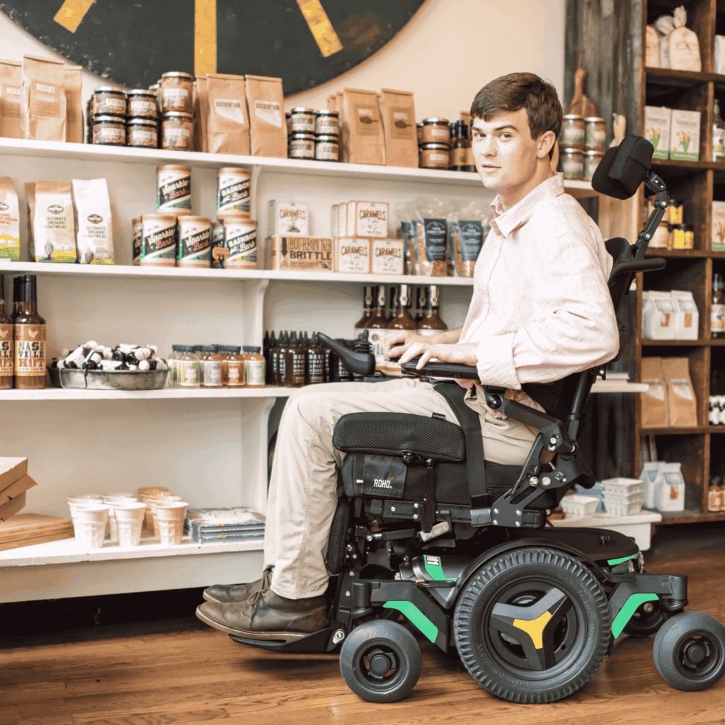 A young man in a power wheelchair browses a small shop filled with gourmet snacks, sauces, and packaged goods. He wears a light pink shirt and khaki pants, and the store has a warm, rustic interior.