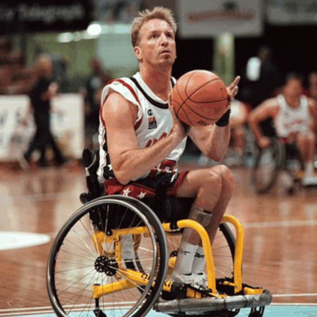 A male adaptive basketball player in action on the court holds a basketball and prepares to shoot during a game, wearing a sports uniform and surrounded by other athletes in wheelchairs.