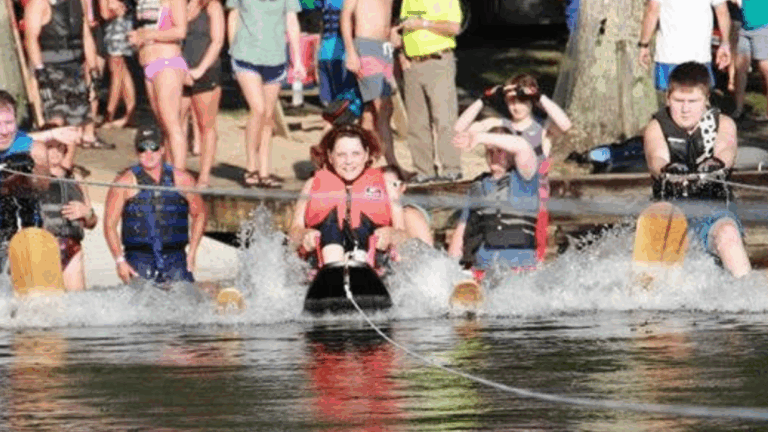A group of people wearing life jackets and water skis, part of Cyndis Story, are being pulled across a lake, splashing water as a crowd watches from the shore in the background.