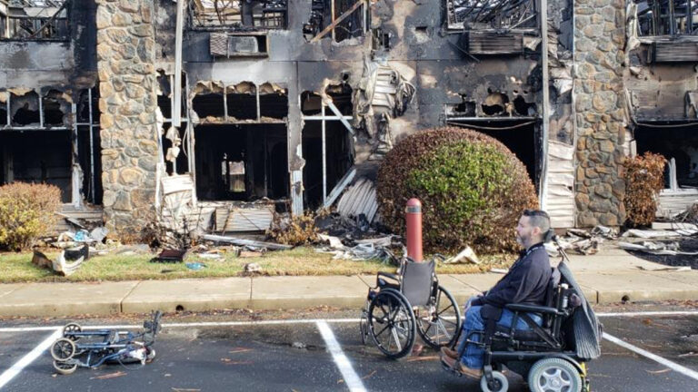 A man in a motorized wheelchair sits in a parking lot in front of a heavily burned building. Nearby, an empty wheelchair and bicycle lie among scattered bushes and debris, hinting at the hardships within Vances Story.