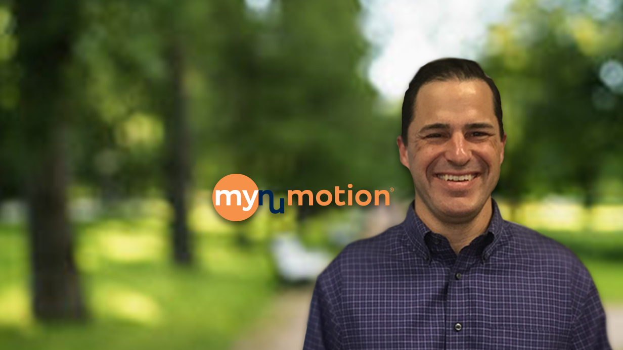 A smiling man in a purple checkered shirt stands in front of a blurred green outdoor background with the mynumotion logo next to him, sharing Mikes Story.