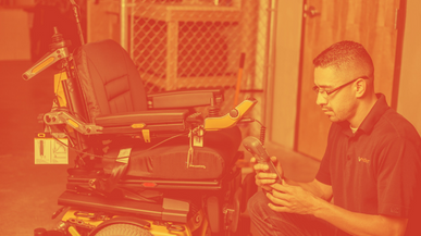 A man kneels next to a motorized wheelchair, examining a handheld device—possibly performing maintenance or repairs for better service by Numotion in an indoor setting.