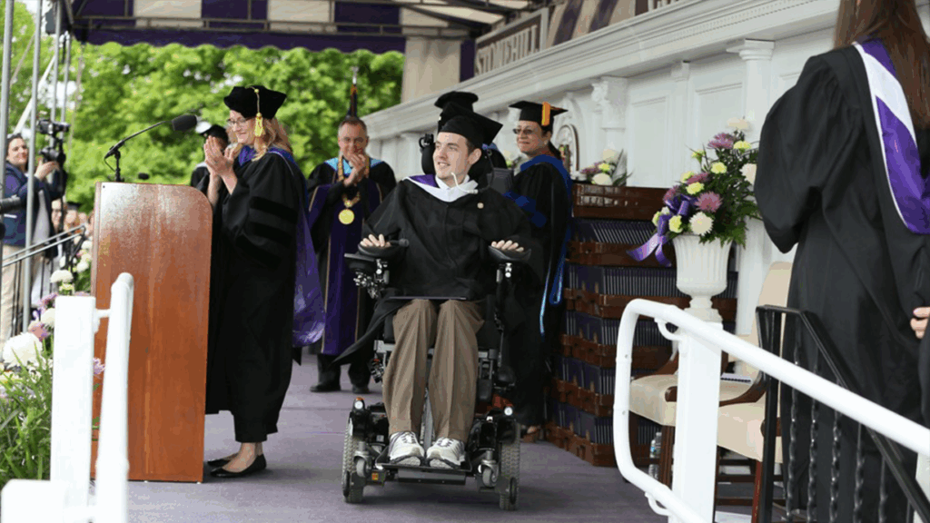 A graduate, part of Matts Story, smiles on stage at a commencement ceremony, surrounded by faculty in academic regalia. A woman at the podium claps as other faculty members stand in the background, also applauding.