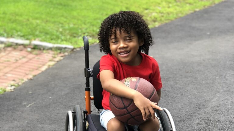 Aydins Story: A young child with curly hair, wearing a red shirt and shorts, smiles while holding a basketball and sitting in a wheelchair on a paved path outdoors. Grass and bricks are visible in the background.