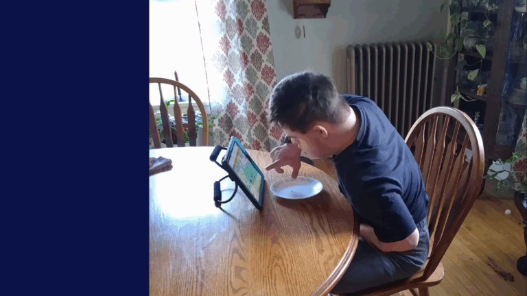 A boy sits at a wooden table, using a tablet propped on a stand with one hand while eating from a white plate with the other. Sunlight streams through patterned curtains—just another moment in Calebs Story.