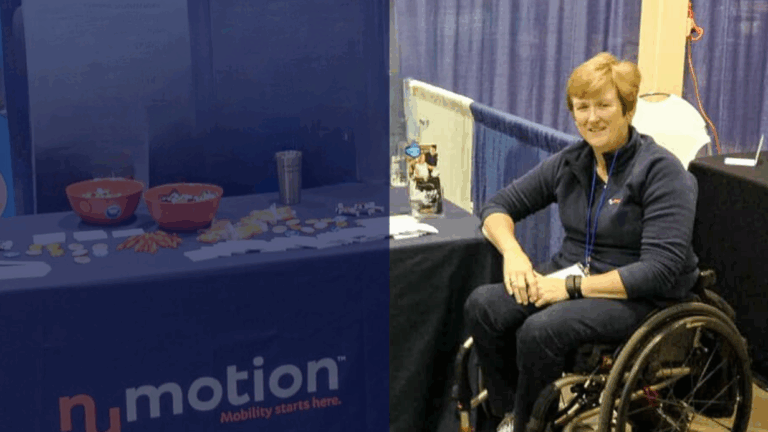 A woman in a wheelchair sits at a booth with a blue tablecloth that reads nmotion Mobility starts here, sharing Carols Story. The table displays bowls, brochures, and small items. Blue curtains hang in the background.