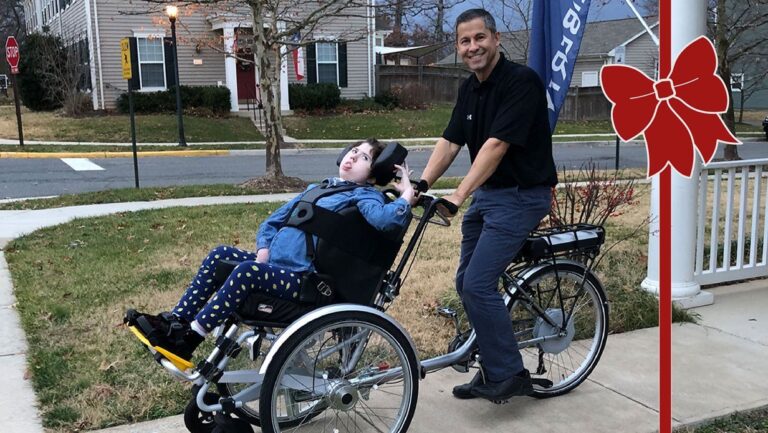 A man smiles while pushing a young person in a specialized wheelchair attached to a bicycle on a suburban sidewalk, sharing joy reminiscent of Carolines Story. A red bow graphic is overlaid on the right side of the image.