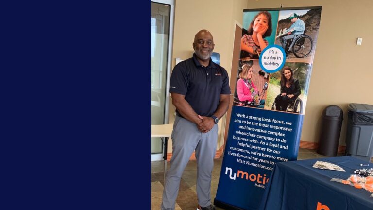 A smiling man stands next to a NuMotion display table featuring brochures, treats, and a banner showing people with disabilities, including a young girl in a wheelchair and a boy with a prosthetic leg—part of Chris’s Story.
