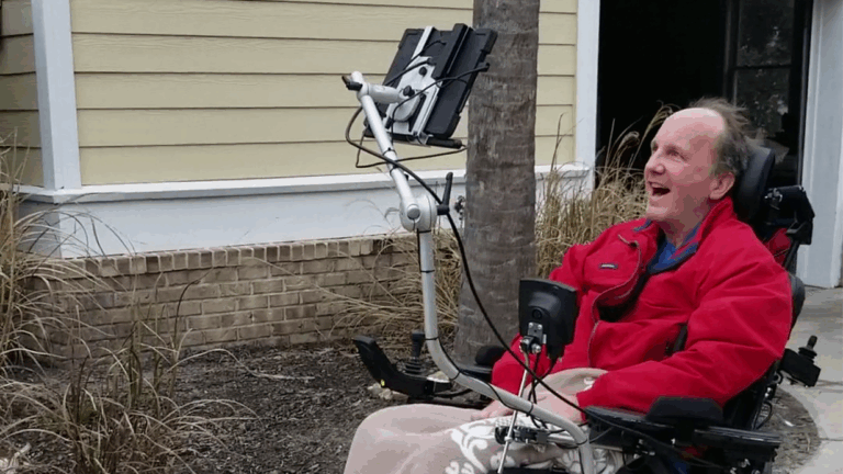 A man in a red jacket sits in a wheelchair outdoors, smiling. He uses assistive technology attached to his wheelchair with a flexible arm. A beige building and some dry grass are visible in the background.