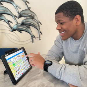 A young man smiles while using a communication app on a tablet with colorful symbol-based buttons. He is seated at a table with a metal dolphin wall sculpture behind him.