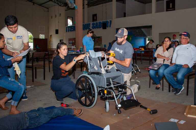 Several people work together to assemble a wheelchair in a community center, inspired by Kriss Story, while others watch and observe from the background. Tools and wheelchair parts are scattered on the floor around them.