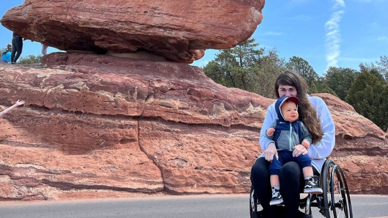 Elizabeths Story: A woman in a wheelchair holds a small child on her lap, both smiling, in front of a large, flat-topped red rock formation outdoors with trees and sky in the background.