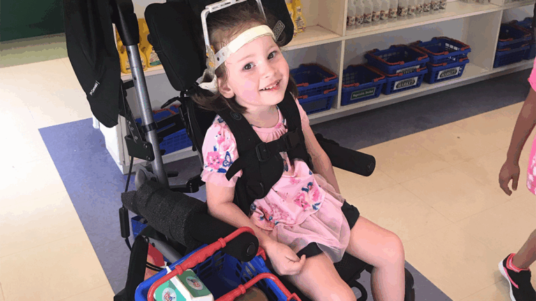Elizabeths Story: A young girl in a pink floral dress and headband smiles at the camera from her wheelchair. Shelves with blue baskets and supplies are visible in the background.