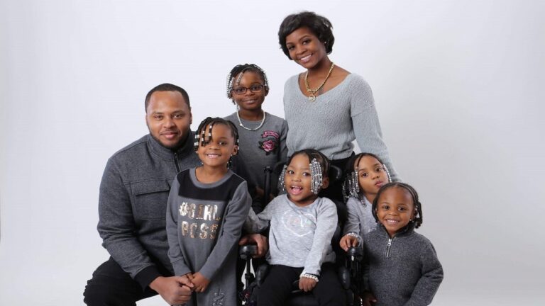A smiling family of six, including two adults and four young girls, poses together against a plain white background. One girl is seated in a wheelchair. They are all wearing coordinated gray outfits, sharing Kiearas Story of unity and joy.
