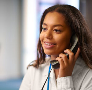 A woman in an office setting speaks on a landline phone. She wears a white blouse and lanyard, smiling as she engages in conversation.