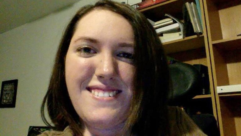 Gabriellas Story: A woman with straight brown hair smiles at the camera. She is indoors, with a bookshelf filled with books and other items in the background.