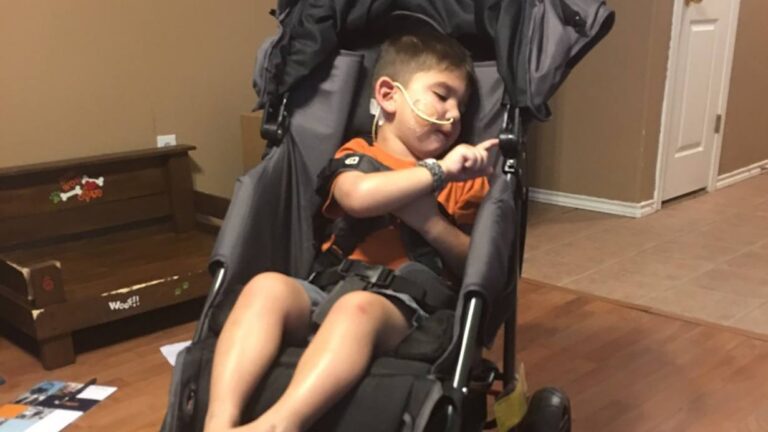 A young boy named Garon with a nasal cannula sleeps in a large stroller indoors. He wears an orange shirt and wristband, his head resting to the side, right arm raised as he touches the stroller’s strap—a quiet moment in his story.