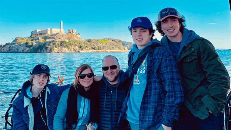 A group of five people, including two adults and three teenagers, pose together on a boat with Alcatraz Island visible in the background on a sunny day. This moment is part of Gregs Story, and they are all dressed in casual, warm clothing.