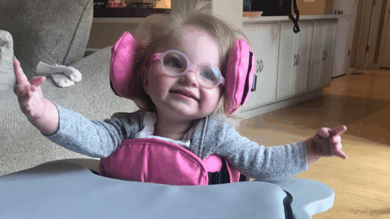 A young child with light hair, wearing glasses and a gray shirt, smiles with arms outstretched while sitting in a pink adaptive chair at a table in a cozy home setting—part of Hatties Story.