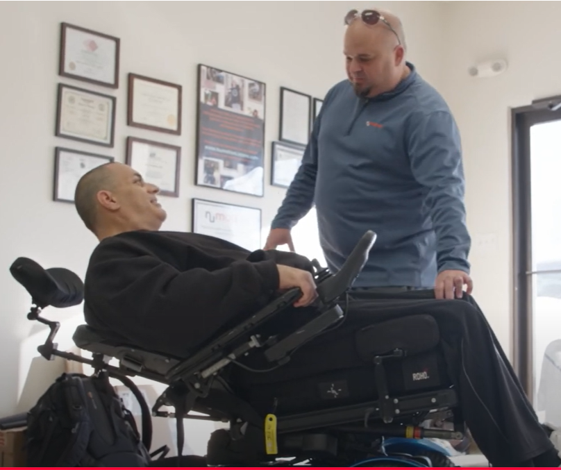 A man in a reclining wheelchair smiles and talks to another man standing beside him in an office with framed certificates on the wall, showcasing the comfort and versatility of complex wheelchairs.