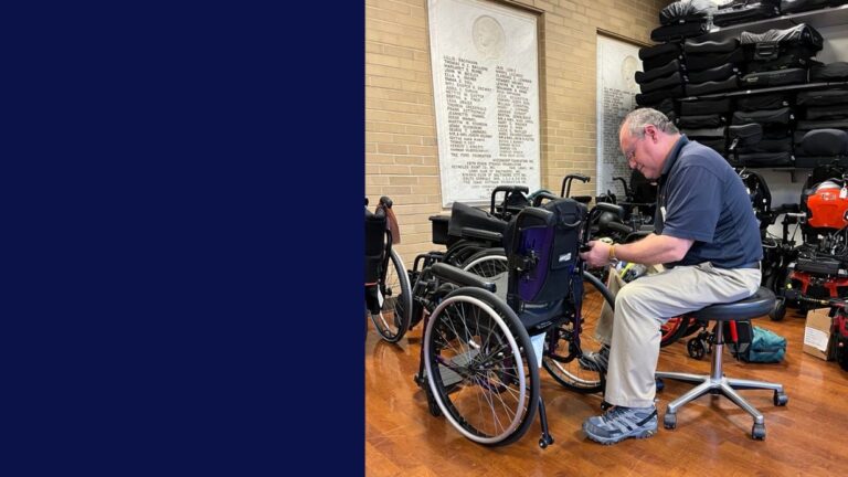 A man sits on a rolling chair indoors, repairing or adjusting a wheelchair. Several wheelchairs and mobility equipment surround him, with plaques on a brick wall in the background—this moment is part of Jeffs Story.