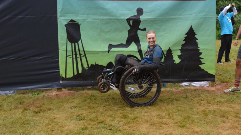 A smiling person in a wheelchair, wearing athletic gear, poses in front of a mural with silhouettes of a runner, water tower, and trees on a grassy area, representing Jillians Story.