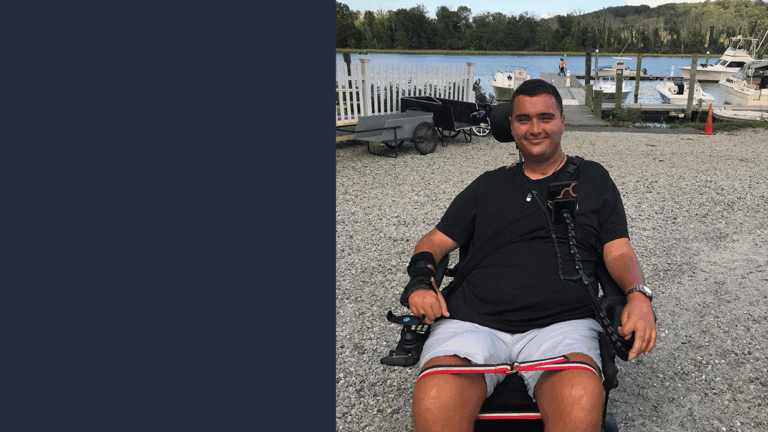 A young man in a motorized wheelchair smiles at the camera near a marina with boats, water, trees, and hills in the background on a sunny day.