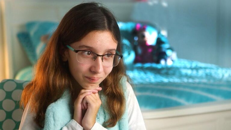 A young girl with long brown hair and glasses sits with her hands clasped under her chin, smiling softly. She wears a light blue shawl in a cozy, blue-themed bedroom, capturing a quiet moment from Kierras Story.