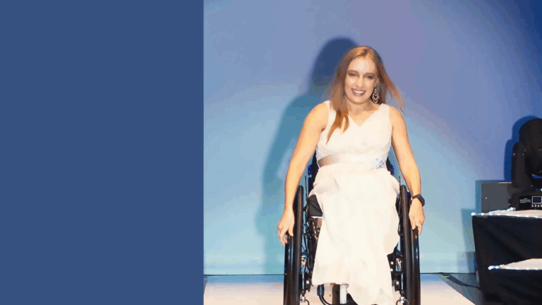 A smiling woman in a white dress sits in a wheelchair on a stage with blue lighting, sharing Kyanns Story.