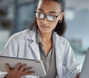 A female healthcare professional in a lab coat reviews documents on a tablet while working at a desk with a laptop. She wears glasses and appears focused.