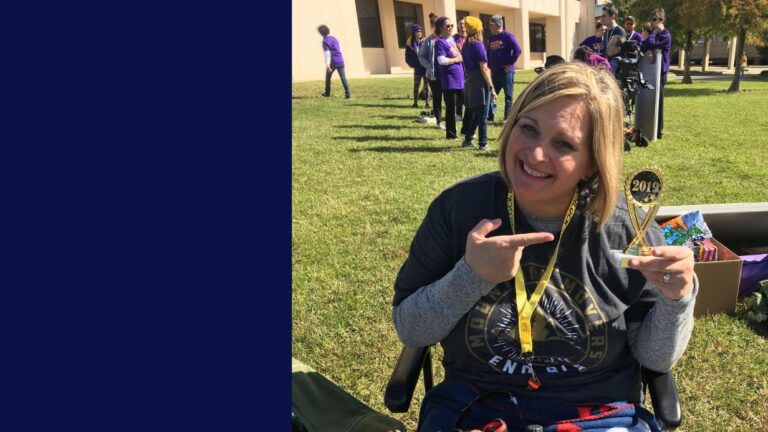 A smiling woman sits on the grass, pointing to a small 2019 trophy she is holding. This moment from Moniques Story features people in purple shirts standing in the background near a building on a sunny day.