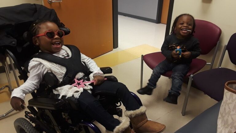 Two young children are laughing together indoors in Kanias Story. One girl, wearing heart-shaped sunglasses, a white sweater, jeans, and boots, sits smiling with a boy in dark clothes who holds a toy.