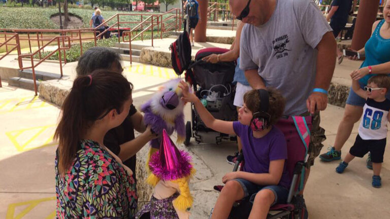 A child in a wheelchair, wearing headphones, smiles and touches a purple puppet held by an adult. Lillies Story unfolds as other adults and children stand nearby outdoors on a sunny day.