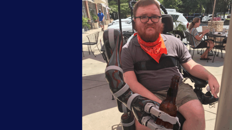 Seans Story: A man in a wheelchair with a robotic arm holds a brown bottle. He wears glasses, a red bandana, and a gray t-shirt, sitting outdoors at a café with people and trees in the background.