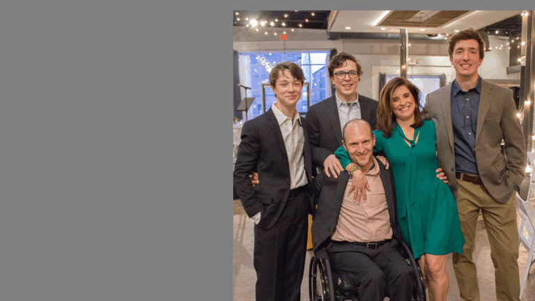 A smiling family of five poses indoors; three young men in suits stand behind, while a woman in a green dress stands beside a man using a wheelchair, all looking at the camera. This moment captures the warmth at the heart of Susans Story.