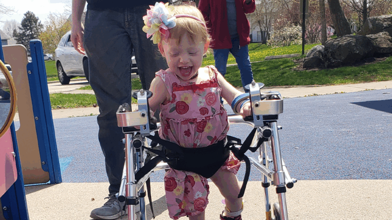 A smiling young girl in a floral dress uses a walker at a playground. Featuring Tessas Story, she wears a flower headband and looks happy and excited, with supportive adults nearby and trees in the background.
