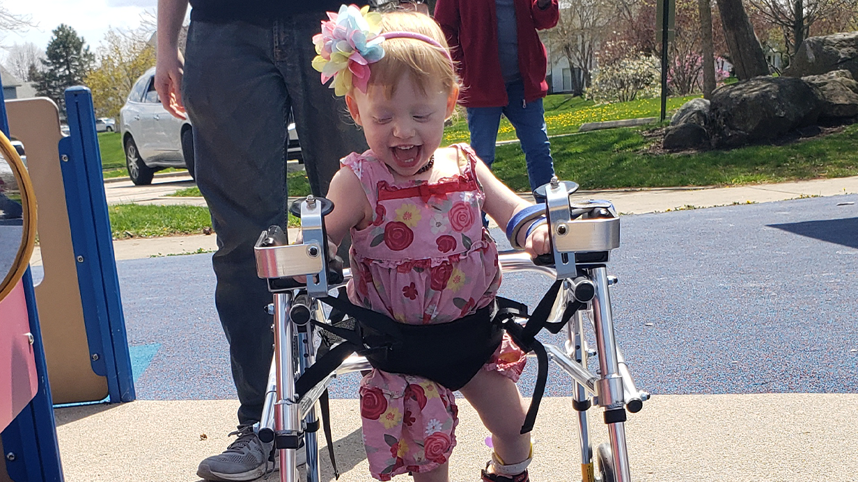 A smiling young girl in a floral dress uses a walker at a playground. Featuring Tessas Story, she wears a flower headband and looks happy and excited, with supportive adults nearby and trees in the background.