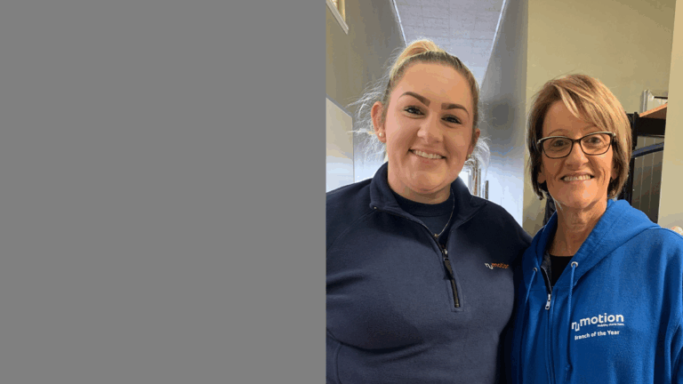 Two women, Tina and Jadi, smile while standing side by side indoors; one wears a dark blue pullover, the other sports glasses and a bright blue nj motion Branch of the Year hoodie. A gray block fills the left part of the image.
