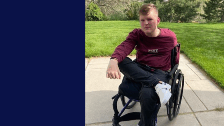 A young man in a maroon long-sleeve shirt uses a wheelchair as he sits on a sunny outdoor patio, green grass and trees behind him. This is part of Porter’s Story.