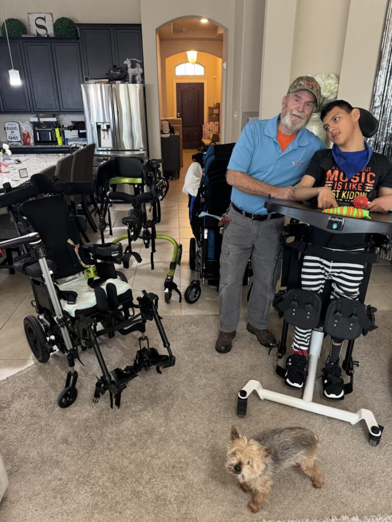 A man in a blue shirt and cap stands next to a smiling boy in a standing frame in a living room, surrounded by several mobility devices awaiting In-Home Repair. A small dog rests nearby, with the kitchen visible in the background.