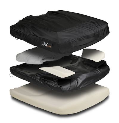 An exploded view of a pediatric wheelchair cushion displaying its black fabric cover, internal foam and padding layers, and white base support, all separated to highlight the seating and positioning structure.