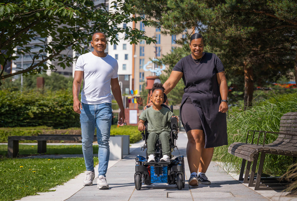 A smiling child in a wheelchair is pushed by a woman while a man walks beside them in a sunny park with trees, grass, benches, and tall buildings in the background—showing what to expect on an inclusive family outing.