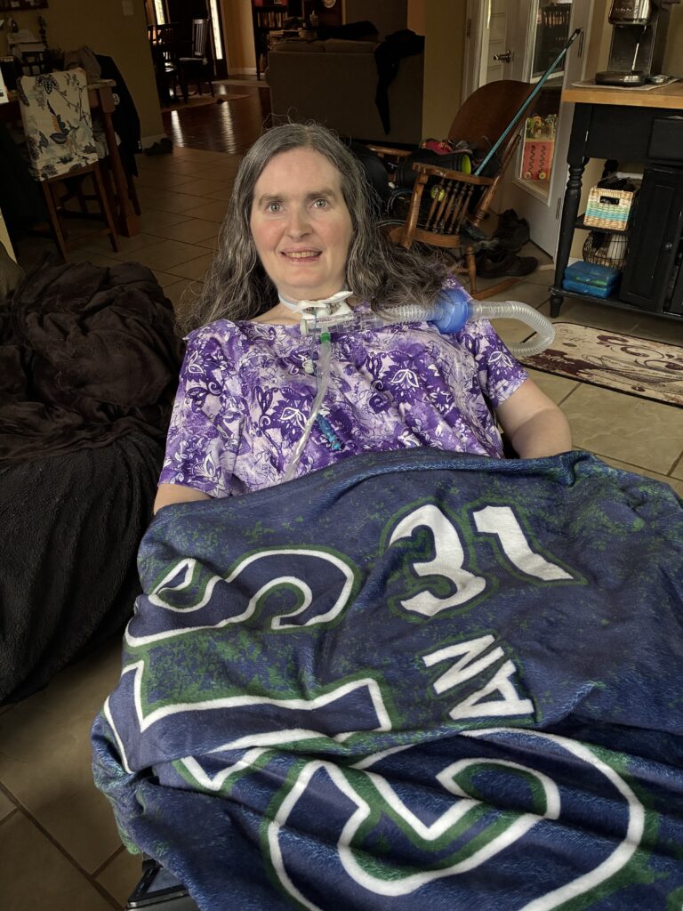 Jessicas Story: A woman with long gray hair, wearing a purple patterned shirt, smiles while sitting in a wheelchair. She is wrapped in a navy blue and green blanket in a cozy, warmly lit living room.