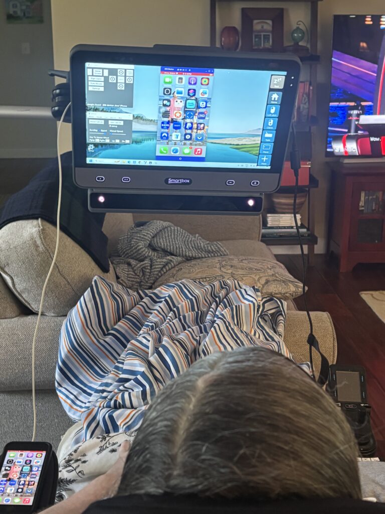 Jessicas Story: A person lying on a couch uses a large communication device displaying various app icons. Covered by a striped blanket, they relax while a TV and other electronics are visible in the background.