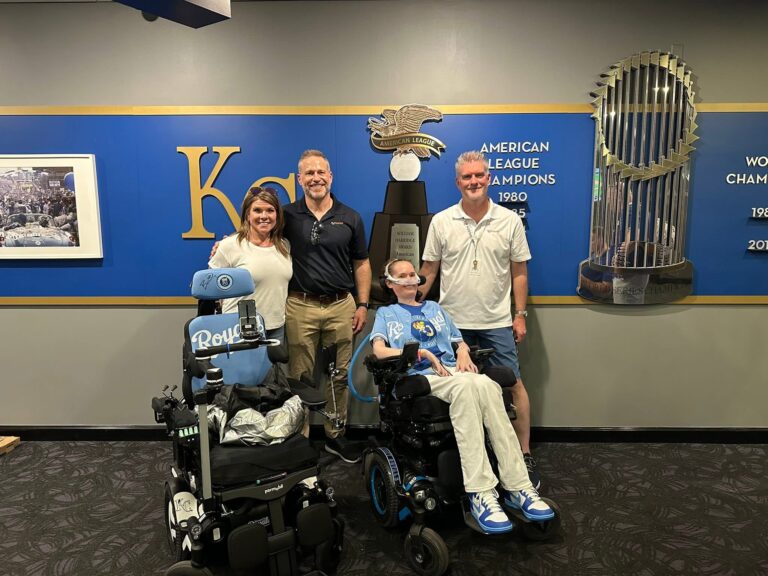 Four people smile for a photo indoors; two are standing, and two are in wheelchairs with Royals gear, sharing Sarahs Story. Kansas City Royals championship displays and a large trophy stand behind them.